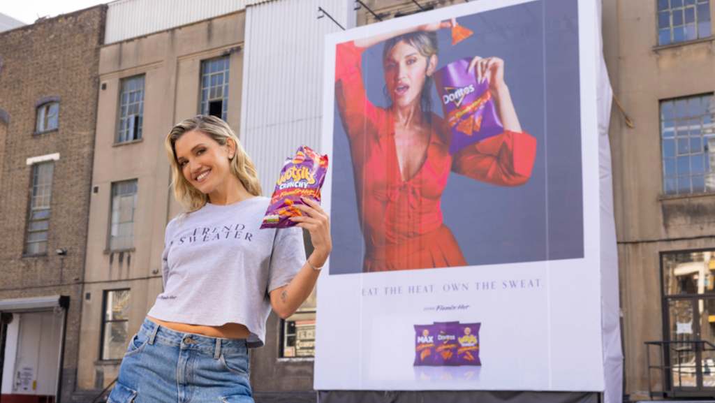 A person stands smiling, holding a bag of spicy snacks in front of a large outdoor advertisement. The ad shows a model eating the same snacks. The setting is urban with brick and industrial buildings.
